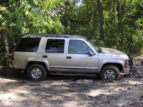 2000 Chevrolet Tahoe - A silver SUV is stuck in a muddy area surrounded by trees.