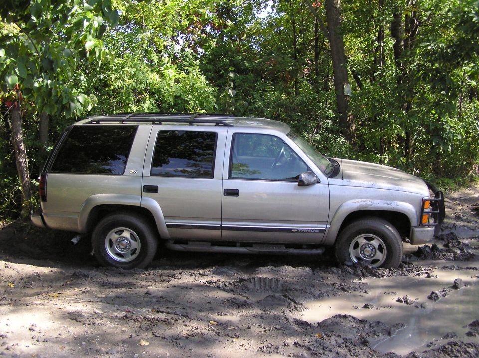 Silver SUV stuck in mud, side view. The vehicle has a roof rack and appears to be from the late 1990s