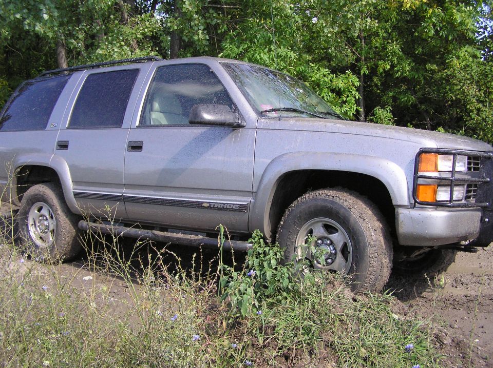 Muddy silver Chevy Tahoe side view with rock rails, brush guard and mud tires