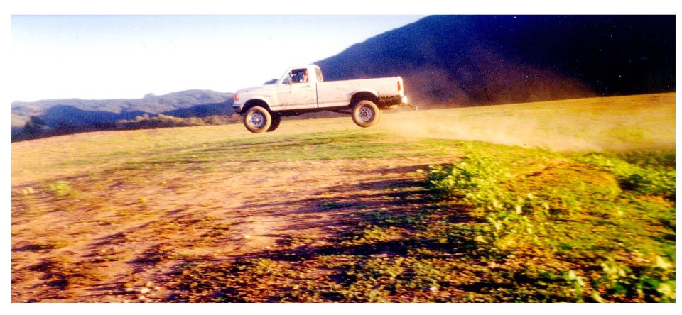 White regular-cab pickup truck airborne mid-jump over a dirt hill with dust trailing behind