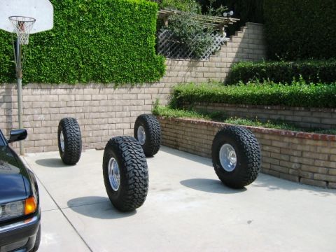 1985 Ford Bronco - Four off-road tires are arranged on a driveway next to a partial view of a black car.