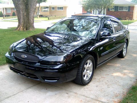2004 Ford Taurus - Black 2004 Ford Taurus sedan parked on a driveway, with tree roots reflected on its hood.