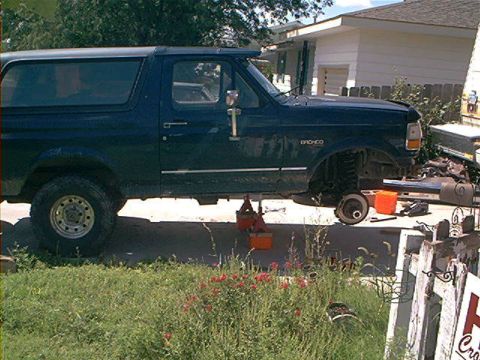 1995 Ford Bronco - A dark blue/black two-door Ford Bronco full-size SUV viewed from the side, raised on jack stands with the front wheel removed exposing the…