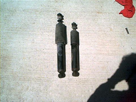1995 Ford Bronco - Two automotive shock absorbers laid side by side on a concrete floor for comparison; the left one has a ribbed dust boot and appears longer…