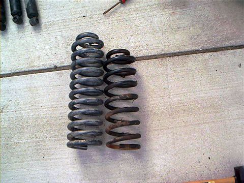 1995 Ford Bronco - Two coil springs laid side by side on a concrete floor, the left one noticeably taller with dark/gray paint and the right one shorter with…