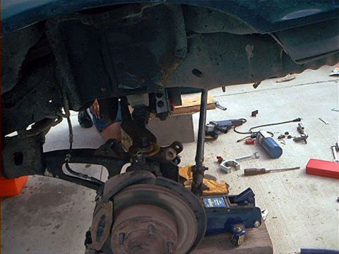 1995 Ford Bronco - Underside view of a dark-colored Ford Bronco raised on a blue floor jack, showing the front suspension, drum/rotor brake assembly, and coil…