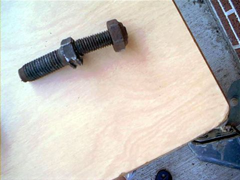 1995 Ford Bronco - A rusty steel bolt with a hex head and a matching hex nut threaded partway down its shank, resting on a light plywood surface. In the backg…