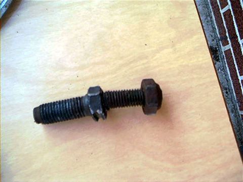 1995 Ford Bronco - A dark, weathered hex-head bolt with a threaded shaft and an additional slotted nut or spacer threaded onto it, resting on a light wood-gra…