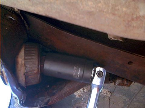 1995 Ford Bronco - Underside view showing a socket on an extension bar and chrome ratchet handle being used on a large bolt of a rusty leaf spring or suspensi…
