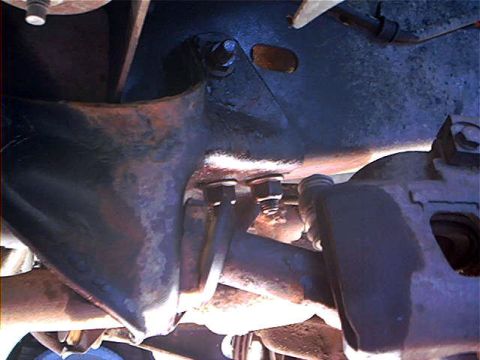 1995 Ford Bronco - Flash-lit view under the truck of rusty front suspension and steering parts: a large stamped bracket bolted up with linkage passing below a…