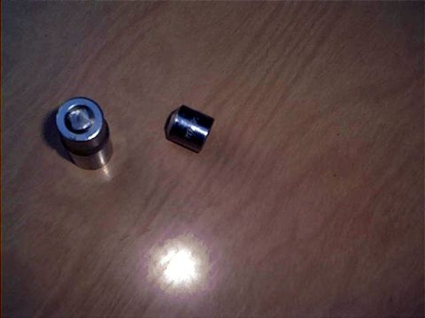 1995 Ford Bronco - Two small metal cylindrical parts sit on a wood-grain surface lit by a bright spotlight; one is a chrome socket-like piece and the other is…