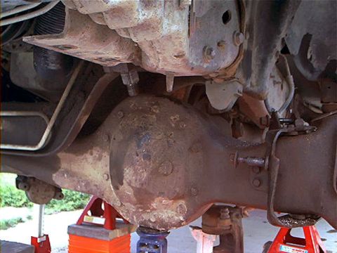 1995 Ford Bronco - Underside view of a Ford Bronco showing the front differential housing, axle, and driveline components, all coated in rust and grime while…