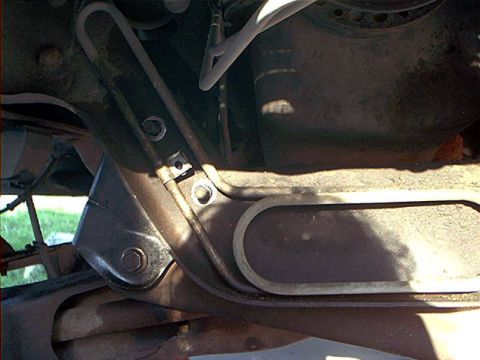 1995 Ford Bronco - Close-up underside view of a vehicle showing a dark metal frame bracket and crossmember, with an oval-shaped fuel tank skid plate or tank b…
