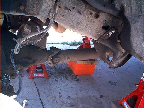 1995 Ford Bronco - Underbody view of a truck on red jack stands, looking along the front driveshaft and U-joint toward the front axle, with brake lines and wi…