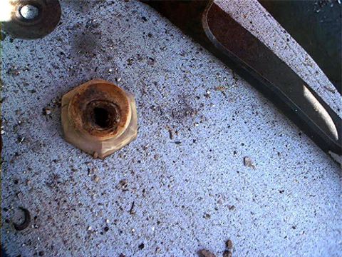 1995 Ford Bronco - Extreme close-up of a rust-caked, rounded-off hex nut with a hollowed threaded center, sitting on a dirty, debris-covered surface with a pr…