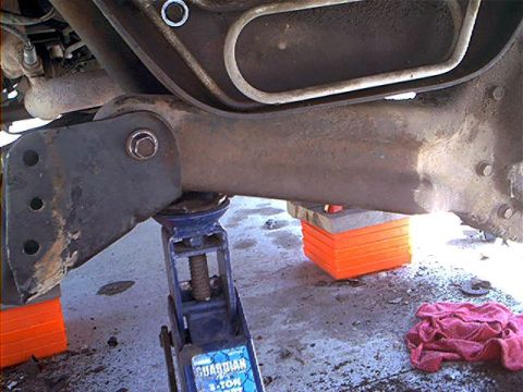 1995 Ford Bronco - Under-vehicle view of front suspension work during a lift install, with a cast axle beam or radius arm resting on a blue bottle jack and a…