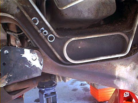1995 Ford Bronco - Underside view of a truck's front crossmember and axle beam area during suspension work, with rust and road grime coating the stamped metal…