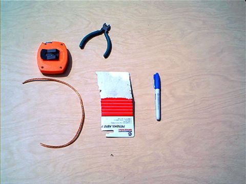 1995 Ford Bronco - Flat-lay on a plywood workbench: an orange tape measure, small wire-cutter pliers, a bent length of braided wire, a blue marker, and a torn…