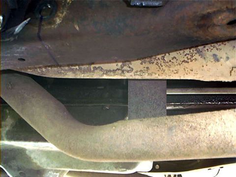 1995 Ford Bronco - Underbody close-up showing an exhaust pipe bending past a black crossmember bar with a rectangular heat shield between them, beneath a surf…