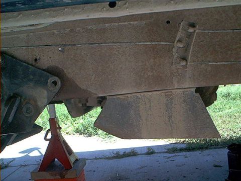 1995 Ford Bronco - Underbody shot looking up at a dusty boxed frame rail and skid plate on a truck supported by a red jack stand on a wood block. Suspension m…