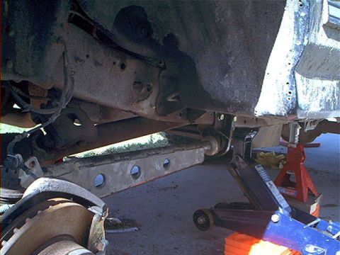 1995 Ford Bronco - Underbody view of a truck raised on a red jack stand and blue floor jack, looking along the boxed frame rail toward a lightweight drilled/l…