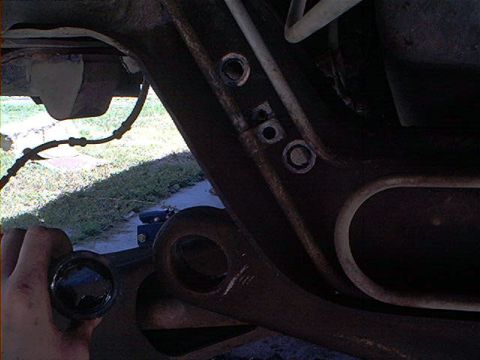 1995 Ford Bronco - Dark under-vehicle shot of a rusty frame rail and crossmember with empty mounting holes and a large lightening slot, taken from ground leve…