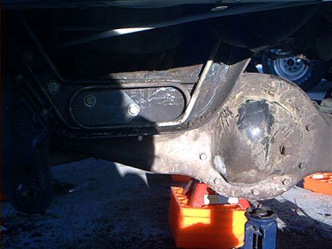 1995 Ford Bronco - Under-vehicle view of a rusty rear axle differential housing with a black suspension bracket bolted above it, photographed in hard sunlight…