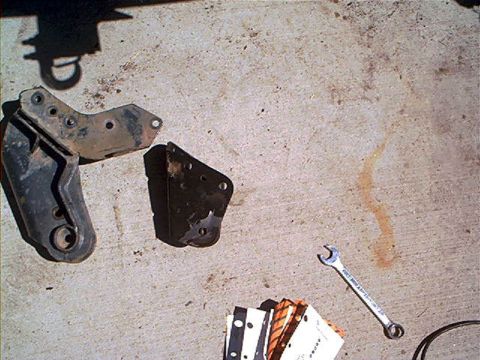 1995 Ford Bronco - Two black steel suspension brackets drilled with multiple mounting holes laid out on sunlit concrete during a lift install, alongside an op…
