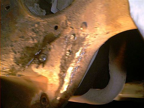 1995 Ford Bronco - Blurry close-up under a vehicle showing a metal frame or bracket section with a rough weld bead and several bolt heads, photographed in war…