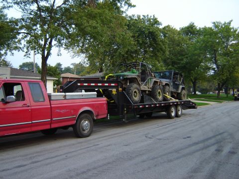 1992 Ford Bronco - A red pickup truck tows a flatbed trailer loaded with two off-road vehicles down a suburban street.