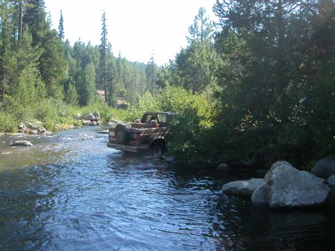 1990 Ford Bronco - A Ford Bronco navigates a rocky riverbed surrounded by dense forest.