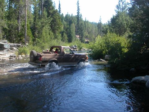 1990 Ford Bronco - A brown 1990 Ford Bronco navigates a river, kicking up water as it drives through the scenic, wooded area.