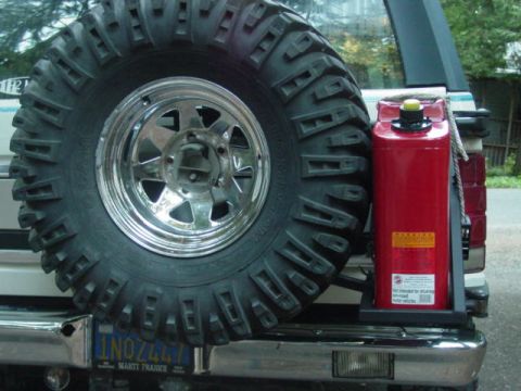 1986 Ford Bronco - Rear-mounted spare tire on a white truck's swing-out carrier: an aggressive mud-terrain tire on a polished slotted aluminum wheel, with a r…