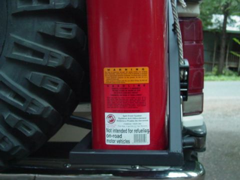 1986 Ford Bronco - Close-up of a red plastic gasoline jerry can mounted in a black steel bracket on a rear tire carrier, with its warning and "Not intended fo…