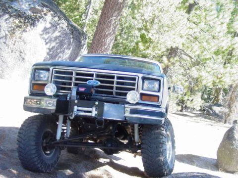 1986 Ford Bronco - Low front view of a lifted blue Ford truck on a granite ledge, showing the chrome eggcrate grille with blue Ford oval, rectangular headligh…