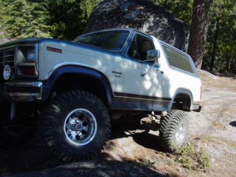 1986 Ford Bronco - Blue-and-white two-tone full-size Ford Bronco flexing over a granite slab in a pine forest, photographed from the driver's-side front three…