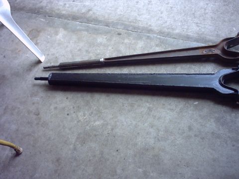1995 Ford Bronco - Two forked suspension radius arms lying side by side on a concrete garage floor — one in bare rusty steel and one boxed and painted black —…