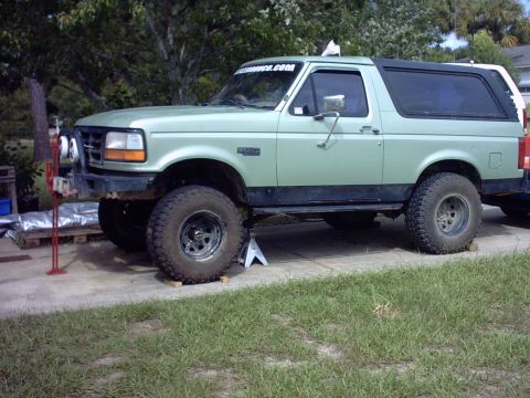 1995 Ford Bronco - Light sage-green full-size Ford Bronco supported on wood blocks and a jack stand on a concrete pad, front three-quarter driver-side view, w…
