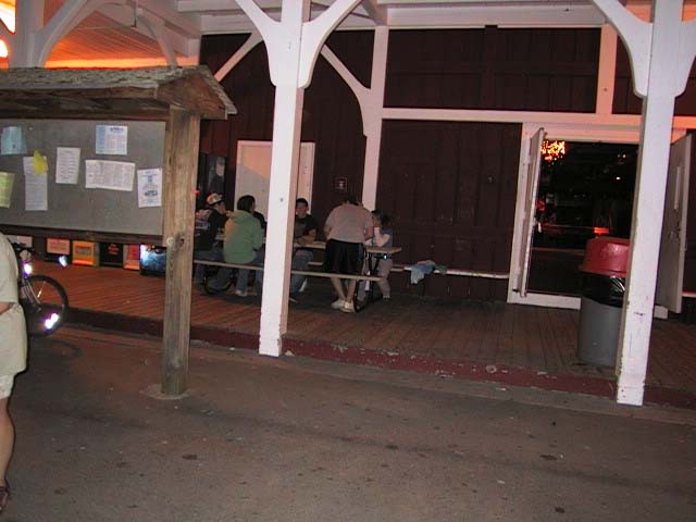 People sit at picnic tables on a wooden deck outside a building with open doors