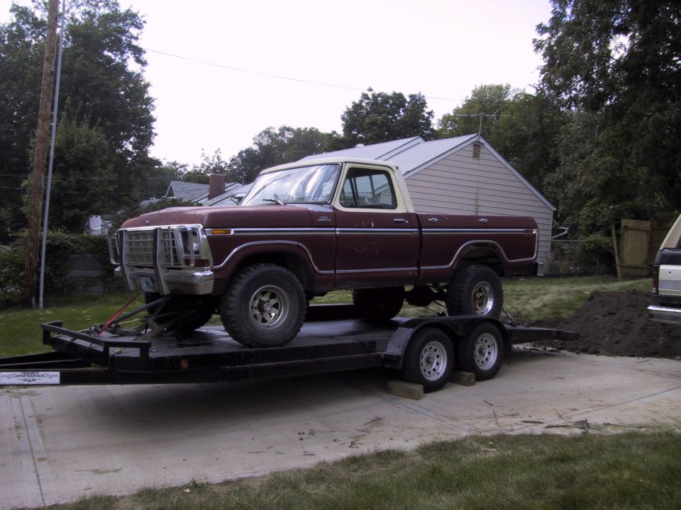 Maroon 1970s Ford pickup with white roof and grille guard chained to a flatbed trailer