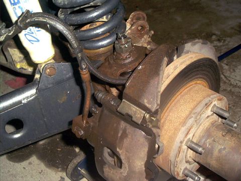 1995 Ford Bronco - Wheel-off view of a solid front axle end during suspension work: a surface-rusted brake rotor and caliper still hang on the knuckle, the fl…