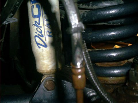 1995 Ford Bronco - Close-up under the front suspension showing a white Dick Cepek shock absorber mounted beside a black coil spring, with a braided stainless…