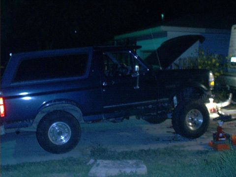 1995 Ford Bronco - A dark blue Ford Bronco full-size SUV with a hardtop shown from the driver's side at night, hood raised and open, with chrome wheels and th…
