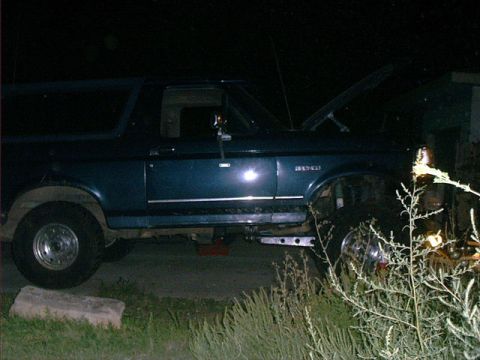 1995 Ford Bronco - Night shot of a dark blue two-door Ford Bronco in profile with the hood open and the right front corner jacked up on stands, front wheel re…