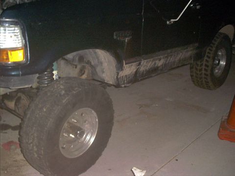 1995 Ford Bronco - Low-angle garage shot of a lifted dark green full-size Ford Bronco/truck, driver-side front wheel on a polished aluminum rim and chunky mud…