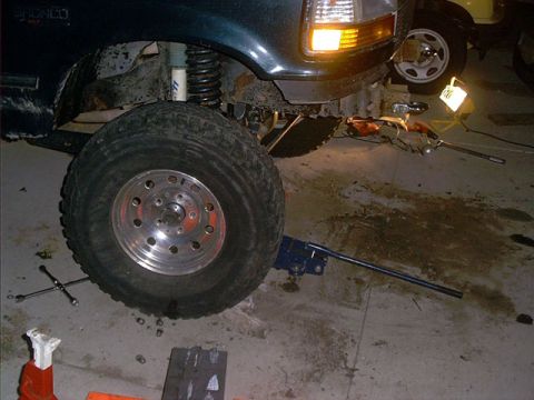 1995 Ford Bronco - Front driver-side corner of a dark green Ford Bronco raised in a garage during suspension work, with a coil spring and radius arm exposed a…