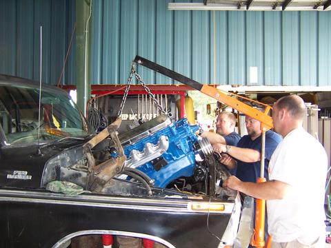 1979 Ford F-150 - An engine is being lowered into the engine bay of a black truck with the help of an engine hoist and several people.
