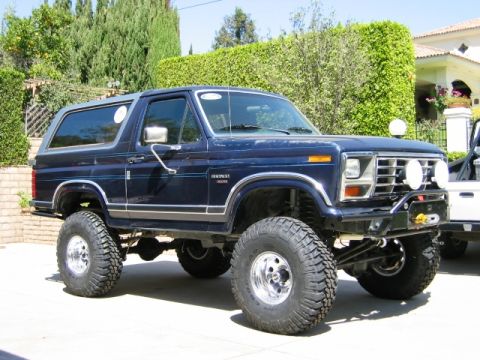 1985 Ford Bronco - A lifted dark blue Ford Bronco with aftermarket lights sits parked in front of a manicured hedge and house.
