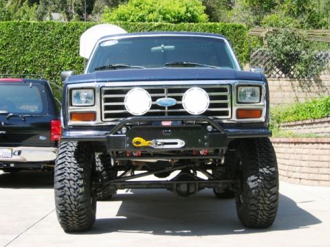 1985 Ford Bronco - Lifted blue Ford Bronco with winch and off-road lights, parked next to a black SUV.