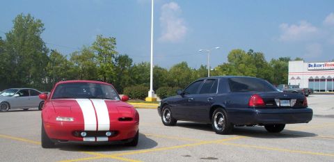 1998 Ford Crown Victoria - A dark blue sedan and a red sports car are parked in a lot near a building.